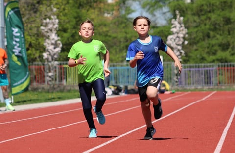 Two young runners competing on a red outdoor track, one wearing a lime green shirt and one in blue, with trees in the background
