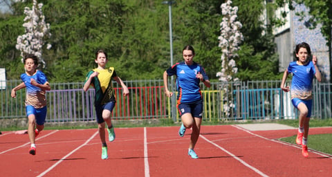 Four young runners sprint on a red outdoor track with white lane lines, wearing athletic gear against a backdrop of green trees and buildings