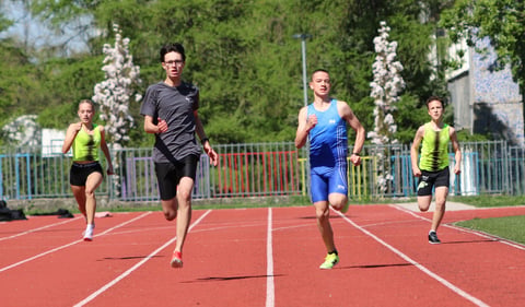 Group of young athletes running on a red outdoor track with green trees and buildings in the background