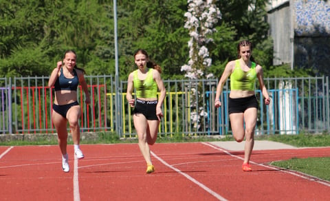 Three female sprinters running on a red outdoor track during a competitive race, with trees and fencing in the background
