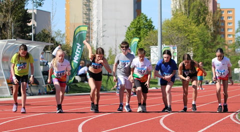 Group of young athletes running on a red outdoor track with city buildings and green trees in the background during a track and field event
