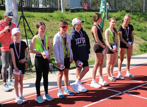 Group of young athletes standing on a red track at an outdoor sporting event with spectators and camera equipment visible