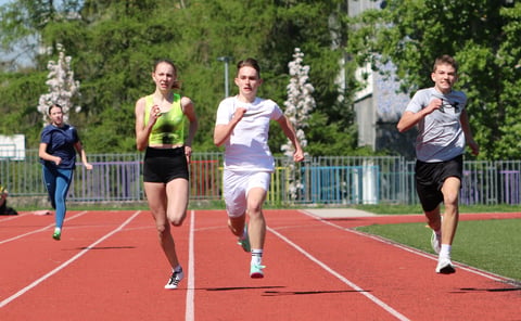 Group of young runners sprinting on a red outdoor track with trees and fencing in the background