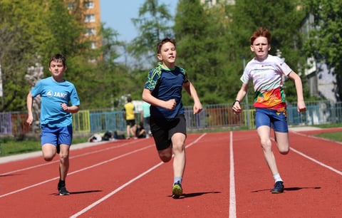 Three young boys running on a red track in a sunny outdoor sports field with trees and buildings in the background