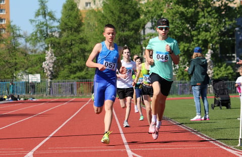 Male runners racing on a red track during a track and field competition, with spectators and buildings in the background