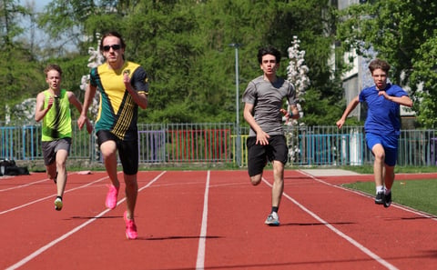 Group of five runners racing on a red outdoor track on a sunny day with trees and fencing in the background