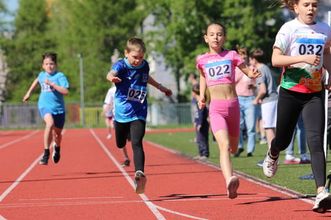 Children running on a red track during a youth sports competition, wearing numbered bibs