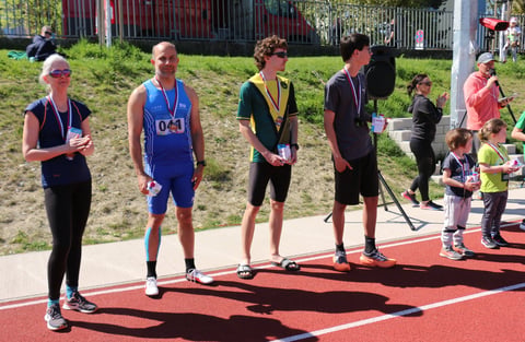 Athletes standing on a red track at an outdoor sporting event, wearing medals and competition bibs, with spectators in the background