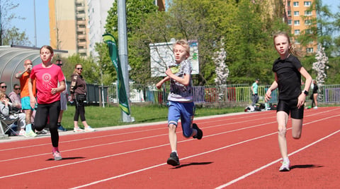 Children running on a red athletic track during a track and field event, with apartment buildings and spectators visible in the background
