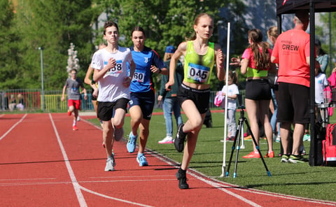 Young runners competing in a track race on a red outdoor track with spectators and event crew members standing nearby