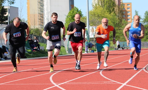 Five male runners wearing race bibs sprint on a red outdoor track with urban buildings and trees in the background