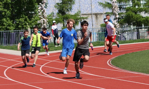 Young athletes running on a red outdoor track during a sunny day with spectators and buildings in the background