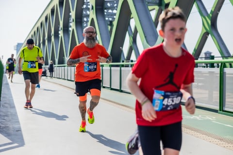 Maratónski bežci prechádzajúci zeleným kovovým mostom počas pretekov