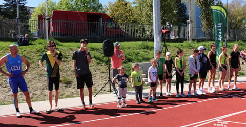 Children lined up on a red track during an outdoor sports class or event with playground and trees visible in background