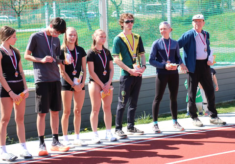 Young athletes and coaches stand together on a sports court at an outdoor tennis facility, wearing matching team apparel and lanyards